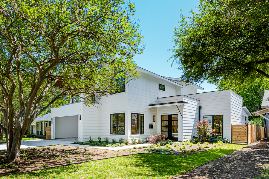 Front view of a modern farmhouse-style home with white horizontal siding, a two-car garage, a covered entry with a black door, and lush landscaping shaded by mature oak trees.