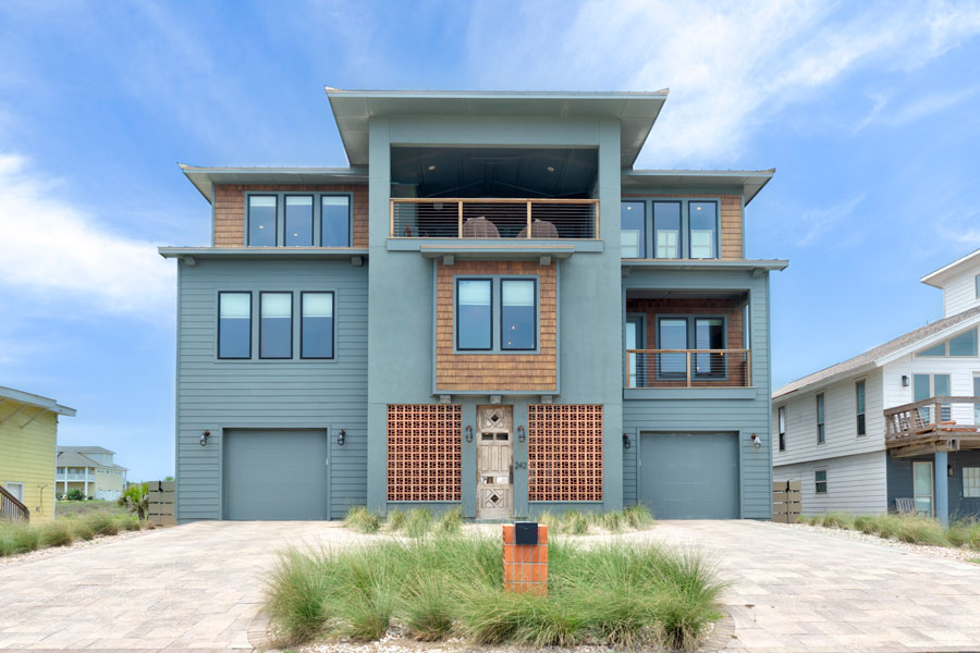 Front elevation of a three-story coastal beach house with blue-gray siding and wood accents, ground-level garage, and a paver driveway landscaped with dune grass.