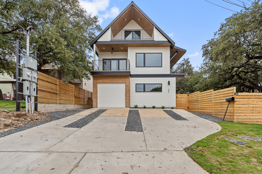 Front exterior of a three-story modern home with a steep gabled roofline, wood accent siding, an upper balcony with metal railing, a two-car garage, and a wood privacy fence beside a permeable paver driveway.