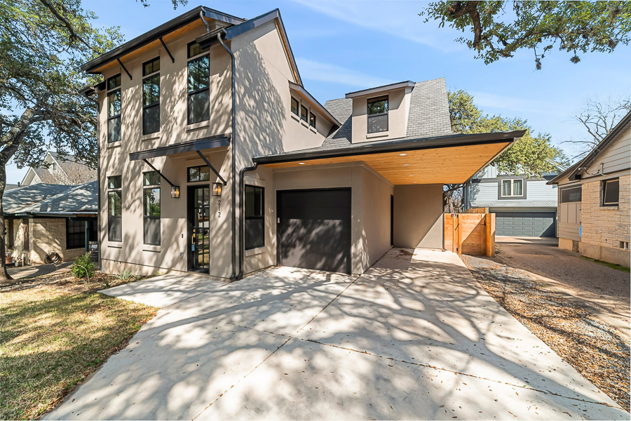 Modern two-story home with tan stucco siding, a dark garage door, an angled wood-lined carport overhang, a dormer window, and a concrete driveway shaded by trees.