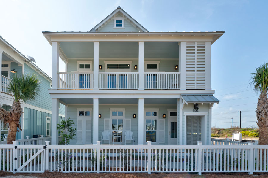 Front elevation of a two-story pale blue coastal cottage with stacked covered porches, white railings, flanking palm trees, and a white picket fence with a gate.