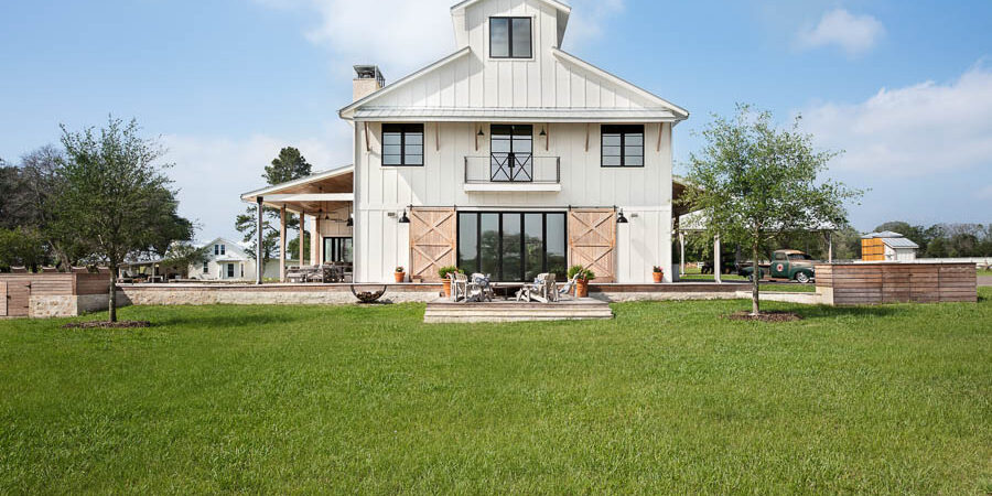 Front elevation of a white farmhouse-style home with a gabled roof, French balcony doors, a covered porch, and a broad front lawn.