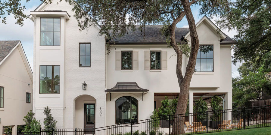 White brick two-story home exterior with steep gabled rooflines, an arched entry, black metal fencing, a mature shade tree in the front yard, and a covered porch with seating.