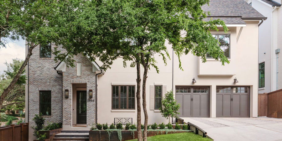 Front elevation of a two-story brick and stucco home with paneled garage doors, a landscaped entry with steps, mature trees, and a paved driveway.