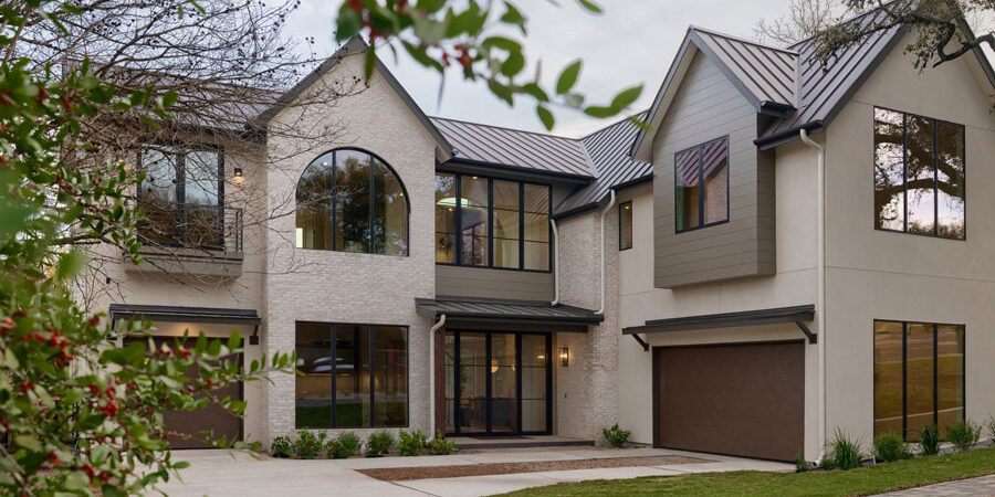 Front exterior of a modern farmhouse home with steep metal roof gables, mixed stucco and wood siding, an arched window, brown garage doors, and a paver driveway with landscaping.