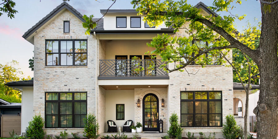 Two-story stone home exterior with black window frames, arched front door, balcony, and tree-lined walkway
