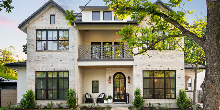 Twilight front elevation of a light brick two-story home with an arched entry door, an upper balcony with decorative railing, black-framed windows, and a landscaped front walkway shaded by a mature tree.